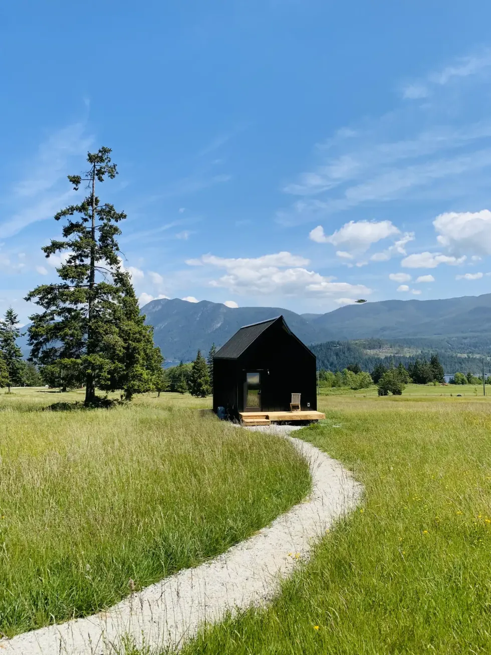 A black cabin sitting in a field of grass with trees and mountains in the distance.